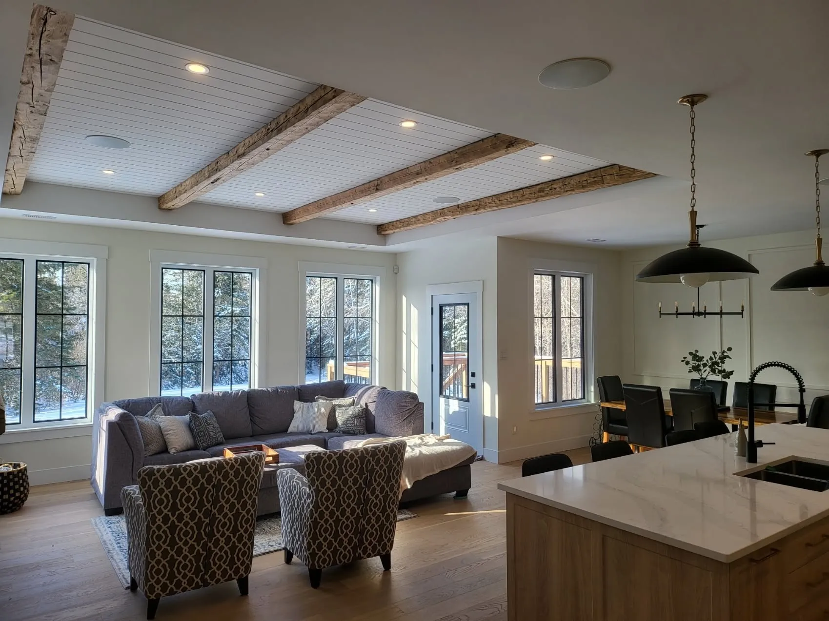 A great room with wood beams across a panelled ceiling, black-framed windows and casing, kitchen island in the foreground
